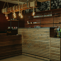 A cozy coffee shop interior featuring a wooden counter with horizontal slats and a well-organized wall with shelves holding various bottles, glasses, and mugs. There is an industrial-style light fixture with exposed bulbs hanging above the bar, adding to the rustic charm. The back wall is decorated with chalkboard-style illustrations and writing.