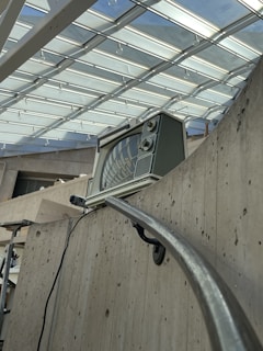 An old-fashioned television set is placed on a concrete balcony railing inside a modern building with a glass ceiling. The ceiling lets in ample natural light, and the reflection of the structure is visible on the television screen. The concrete walls and railing give the space an industrial feel.