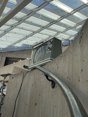 An old-fashioned television set is placed on a concrete balcony railing inside a modern building with a glass ceiling. The ceiling lets in ample natural light, and the reflection of the structure is visible on the television screen. The concrete walls and railing give the space an industrial feel.