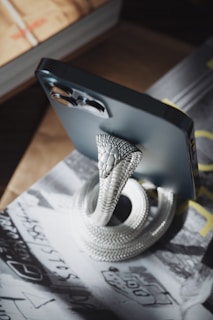 A decorative snake-shaped stand holds up a smartphone. The snake is metallic and intricately designed with a coiled body and detailed scales. The smartphone, in a dark color, is positioned upright, displaying its rear camera module prominently. The setup rests on a surface covered with a magazine or book, featuring black and white text and images.