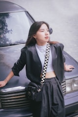 A young woman is leaning against the front of a car. She is dressed in a casual yet stylish outfit featuring a leather jacket, a white top, and high-waisted trousers. A checked strap is connected to a small black shoulder bag she is wearing. Her hair is loose, and she gazes thoughtfully at something off-camera.