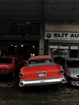 A classic red car is parked in front of an auto repair shop, flanked by two modern vehicles. The auto shop has a sign in both English and another language, featuring a logo similar to BMW. The area appears slightly wet, indicating recent rain.