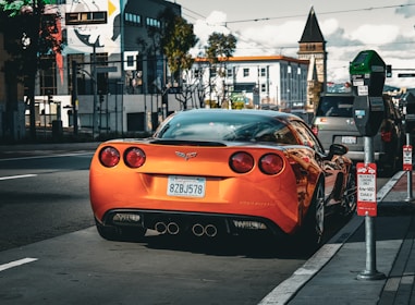 A bright orange sports car is parked on the side of an urban street beside a parking meter. The car is a Corvette, as indicated by the badge and license plate. Surrounding the car are various signs and modern buildings, and a cloudy sky is visible in the background.