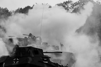 A black and white scene with military tanks partially obscured by thick smoke. Several soldiers are visible among the tanks, and the dense forest in the background creates a dramatic contrast. The scene conveys a sense of action and intensity.