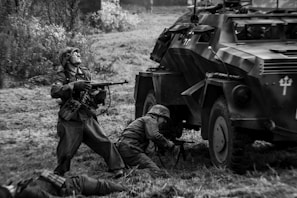 Soldiers in historical military uniforms are engaged in a battle scene next to an armored vehicle. One soldier is standing and aiming a rifle, while another is crouching near the vehicle. The scene is set outdoors in what appears to be a grassy area.