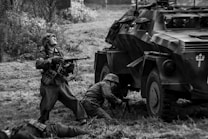 Soldiers in historical military uniforms are engaged in a battle scene next to an armored vehicle. One soldier is standing and aiming a rifle, while another is crouching near the vehicle. The scene is set outdoors in what appears to be a grassy area.