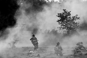 A black and white image of soldiers moving through a misty field, with some taking cover on the ground and others in crouched positions, armed and alert. Vegetation and trees are visible in the background, adding to the tactical setting.