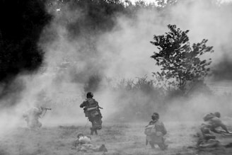 A black and white image of soldiers moving through a misty field, with some taking cover on the ground and others in crouched positions, armed and alert. Vegetation and trees are visible in the background, adding to the tactical setting.