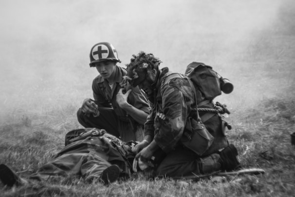 Two individuals in military attire are shown in a smoky, grassy field. One appears to be a medic, recognizable by a helmet with a red cross, kneeling beside another person who is lying on the ground. Another person in full gear, complete with a helmet and backpack, is attending to the individual on the ground. The scene captures a moment of urgency and care.