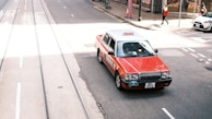 A red and white taxi is driving on a city street with tram tracks visible. The street appears bustling, with a pedestrian in the background and other vehicles parked nearby. The sunlight casts shadows on the street, creating a vibrant urban scene.