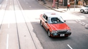 A red and white taxi is driving on a city street with tram tracks visible. The street appears bustling, with a pedestrian in the background and other vehicles parked nearby. The sunlight casts shadows on the street, creating a vibrant urban scene.