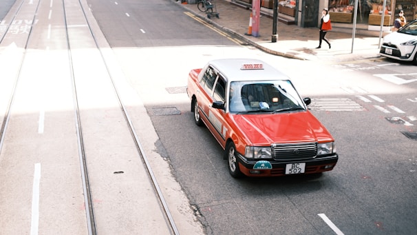 A red and white taxi is driving on a city street with tram tracks visible. The street appears bustling, with a pedestrian in the background and other vehicles parked nearby. The sunlight casts shadows on the street, creating a vibrant urban scene.