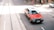 A red and white taxi is driving on a city street with tram tracks visible. The street appears bustling, with a pedestrian in the background and other vehicles parked nearby. The sunlight casts shadows on the street, creating a vibrant urban scene.