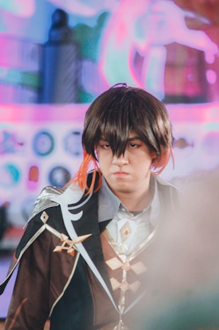 A person dressed in elaborate cosplay attire, featuring a dark brown coat with ornate designs, wearing a wig with brown and orange highlights. The background is blurred with colorful, abstract shapes.