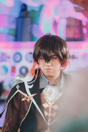 A person dressed in elaborate cosplay attire, featuring a dark brown coat with ornate designs, wearing a wig with brown and orange highlights. The background is blurred with colorful, abstract shapes.