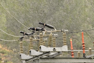 A power line component featuring insulators and cables is set against a backdrop of rain. The rain creates a blurred effect, suggesting a stormy or wet weather condition. The insulators are primarily cylindrical and aligned horizontally, while metal components connect them.