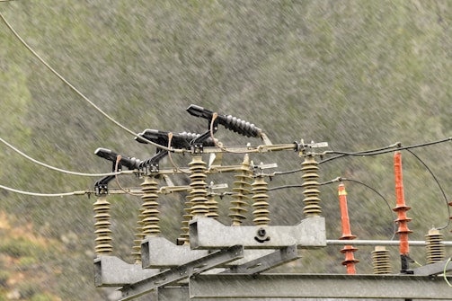 A power line component featuring insulators and cables is set against a backdrop of rain. The rain creates a blurred effect, suggesting a stormy or wet weather condition. The insulators are primarily cylindrical and aligned horizontally, while metal components connect them.