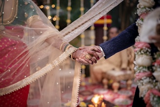 A couple holding hands during a traditional ceremony, with intricate henna designs on the bride's hand and a decorative veil partly obscuring her. The setting includes decorative lights and flowers, indicating a celebratory occasion. The blurred background features a flame, adding a warm glow.