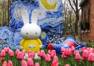 A large white rabbit character wearing a yellow shirt stands in a garden filled with pink tulips. Behind it is a colorful backdrop resembling Van Gogh's 'Starry Night'. A large decorative blue and red clog lies among the flowers. Trees and a brick building can be seen in the background.