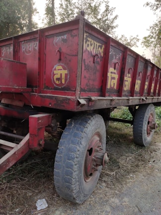 A weathered red trailer with large wheels is parked on a dirt road, surrounded by sparse vegetation. The trailer has worn paint and some text in a non-English script.