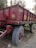 A weathered red trailer with large wheels is parked on a dirt road, surrounded by sparse vegetation. The trailer has worn paint and some text in a non-English script.