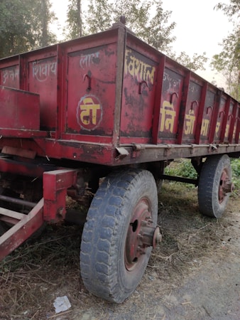 A weathered red trailer with large wheels is parked on a dirt road, surrounded by sparse vegetation. The trailer has worn paint and some text in a non-English script.