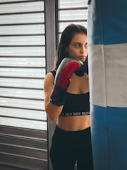 A person wearing a black sports top and red boxing gloves stands next to a blue and white punching bag. The background features horizontal metal shutters, and the person appears focused and contemplative.
