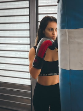 A person wearing a black sports top and red boxing gloves stands next to a blue and white punching bag. The background features horizontal metal shutters, and the person appears focused and contemplative.