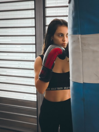 A person wearing a black sports top and red boxing gloves stands next to a blue and white punching bag. The background features horizontal metal shutters, and the person appears focused and contemplative.