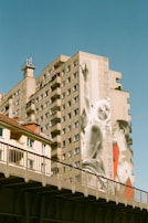 A tall apartment building with a large mural painted on its side, depicting two figures. The mural features one figure gesturing upwards and another figure in red looking towards the first. The building is adjacent to a smaller structure with a tiled roof, and there's a railway bridge in the foreground.