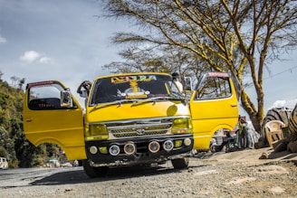 A bright yellow minibus is parked on a rough, unpaved road with its doors open. The vehicle has decorative elements on the windshield and front bumper. Several tree branches and foliage can be seen in the background along with a few scattered clouds in the sky.