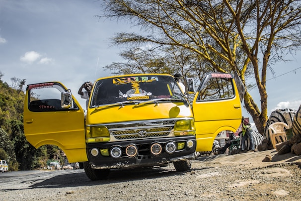A bright yellow minibus is parked on a rough, unpaved road with its doors open. The vehicle has decorative elements on the windshield and front bumper. Several tree branches and foliage can be seen in the background along with a few scattered clouds in the sky.