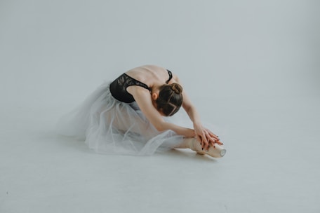 A ballet dancer dressed in a black leotard and a white tulle skirt is gracefully stretching on the floor. She is sitting with her legs extended and bent forward to touch her foot with both hands. Her hair is neatly tied in a bun, and she is positioned on a smooth, light-colored surface.