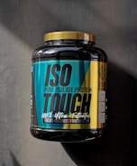 A large black container of ISO TOUCH pure isolate protein with a black and teal label, featuring bold text and nutritional information. The label includes details such as '100% Ultra-Filtrated', 'ISO PURE ISOLATE PROTEIN', and quantities of protein, BCAA, and glutamine. The container has a yellow cap and is placed on a dark textured surface with a striped pattern.