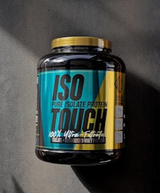 A large black container of ISO TOUCH pure isolate protein with a black and teal label, featuring bold text and nutritional information. The label includes details such as '100% Ultra-Filtrated', 'ISO PURE ISOLATE PROTEIN', and quantities of protein, BCAA, and glutamine. The container has a yellow cap and is placed on a dark textured surface with a striped pattern.