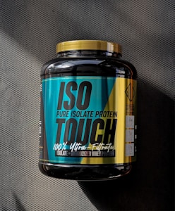 A large black container of ISO TOUCH pure isolate protein with a black and teal label, featuring bold text and nutritional information. The label includes details such as '100% Ultra-Filtrated', 'ISO PURE ISOLATE PROTEIN', and quantities of protein, BCAA, and glutamine. The container has a yellow cap and is placed on a dark textured surface with a striped pattern.