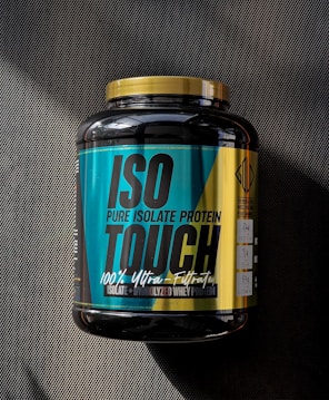 A large black container of ISO TOUCH pure isolate protein with a black and teal label, featuring bold text and nutritional information. The label includes details such as '100% Ultra-Filtrated', 'ISO PURE ISOLATE PROTEIN', and quantities of protein, BCAA, and glutamine. The container has a yellow cap and is placed on a dark textured surface with a striped pattern.