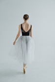 A ballet dancer is captured from behind, standing on pointe. She wears a black leotard with lace detailing on the back and a flowing white tulle skirt. Her hair is styled in a neat bun, and she is poised gracefully in a minimalist, light-filled space.