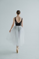 A ballet dancer is captured from behind, standing on pointe. She wears a black leotard with lace detailing on the back and a flowing white tulle skirt. Her hair is styled in a neat bun, and she is poised gracefully in a minimalist, light-filled space.