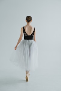 A ballet dancer is captured from behind, standing on pointe. She wears a black leotard with lace detailing on the back and a flowing white tulle skirt. Her hair is styled in a neat bun, and she is poised gracefully in a minimalist, light-filled space.