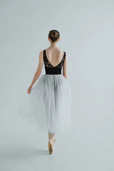 A ballet dancer is captured from behind, standing on pointe. She wears a black leotard with lace detailing on the back and a flowing white tulle skirt. Her hair is styled in a neat bun, and she is poised gracefully in a minimalist, light-filled space.