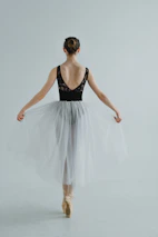 A ballet dancer stands poised on pointe, facing away while wearing a delicate white tutu and a black tank top. The hair is styled in a neat bun, and the background is minimalistic and light gray, emphasizing the elegance and grace of the pose.