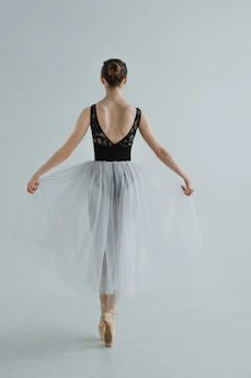 A ballet dancer stands poised on pointe, facing away while wearing a delicate white tutu and a black tank top. The hair is styled in a neat bun, and the background is minimalistic and light gray, emphasizing the elegance and grace of the pose.