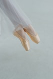 A pair of feet wearing ballet pointe shoes, poised gracefully on tiptoe, are captured against a soft, muted background. The elegant tulle of a ballet costume gently drapes down, emphasizing the delicacy and precision of the pose.