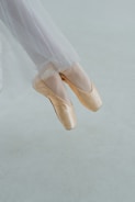 A pair of feet wearing ballet pointe shoes, poised gracefully on tiptoe, are captured against a soft, muted background. The elegant tulle of a ballet costume gently drapes down, emphasizing the delicacy and precision of the pose.