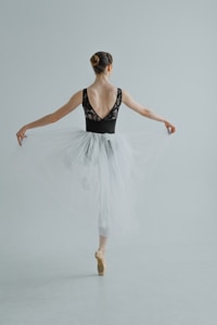 A ballet dancer gracefully stands en pointe, with her back turned. She wears a black leotard with lace accents and a white tulle skirt. Her arms are extended to the sides, and her hair is neatly styled in a bun.