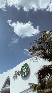 A modern building with a large logo featuring green leaves on its facade. A clear blue sky with a few fluffy clouds is present. Palm tree leaves can be seen in the foreground, adding a tropical atmosphere.