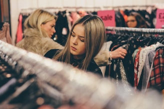 A young woman with long blonde hair is browsing through clothing on a rack in a store. She appears focused on her selection, with sale signs visible in the background. Other shoppers can also be seen looking through clothes.