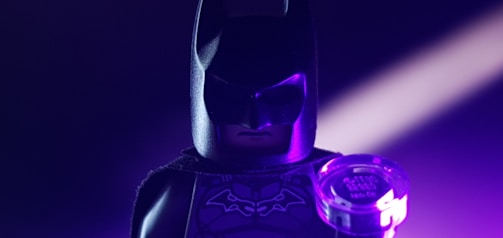 A Lego Batman figure is prominently featured, with dramatic purple lighting creating a moody atmosphere. The figure is holding a small circular object with engraved text, possibly a light source or gadget.