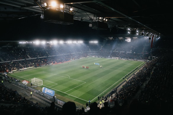 A large illuminated stadium filled with spectators surrounding a green soccer field, where two teams are lined up in the center. Bright stadium lights create a vibrant atmosphere, with banners and advertisements visible around the perimeter.